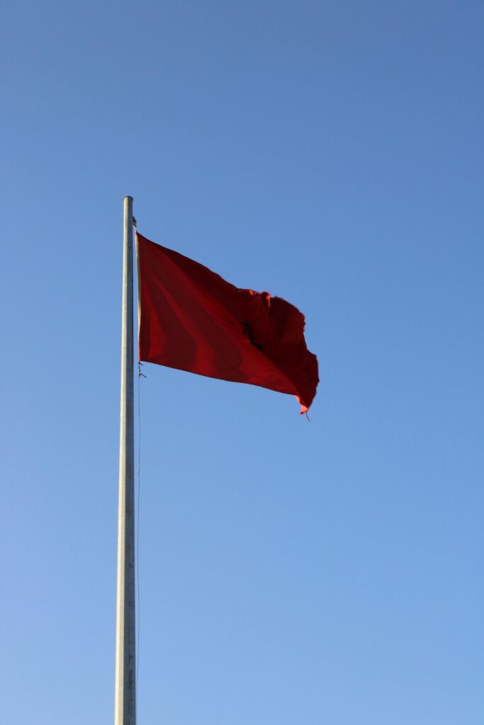 A vibrant red flag flutters in the breeze against a clear blue sky, symbolizing various warning or cultural meanings.